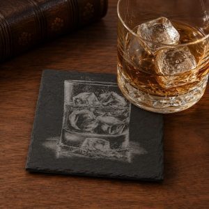 Whiskey tumbler with ice resting on an engraved slate drinks coaster on a walnut table