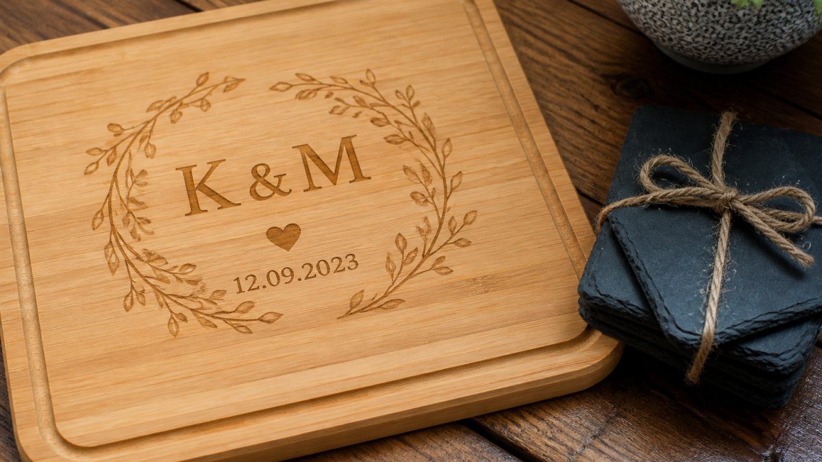 Close-up of a beautifully engraved wooden chopping board with a couple's initials and wedding date, arranged alongside personalised slate coasters on a rustic wooden table with soft natural lighting highlighting the craftsmanship